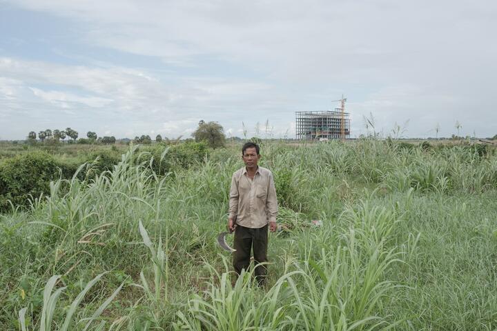 On the outskirts of the city, a farmer poses with his sickle. In the distance, a building is under construction. 